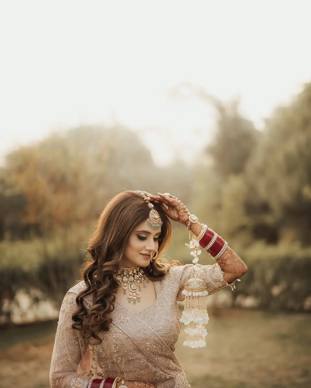 A bride in a cream lehenga with gold jewellery and kaleere at her wrist, one hand raised to her hair, a sunlit garden behind her.