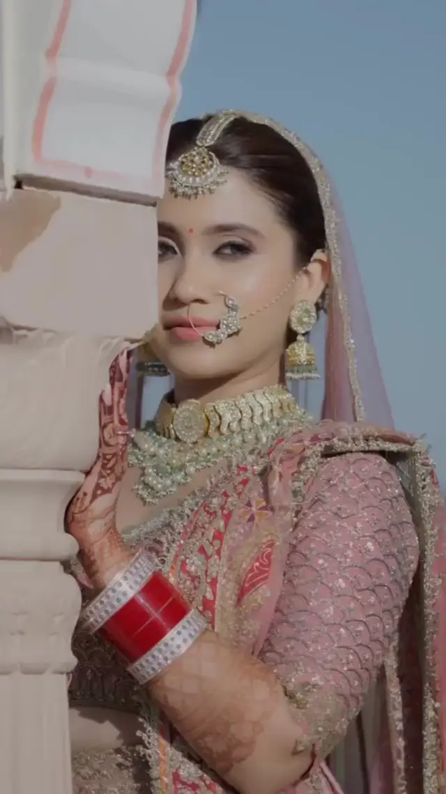 A bride in a pink lehenga with a gold choker and nose ring, standing beside a white pillar under a clear sky.