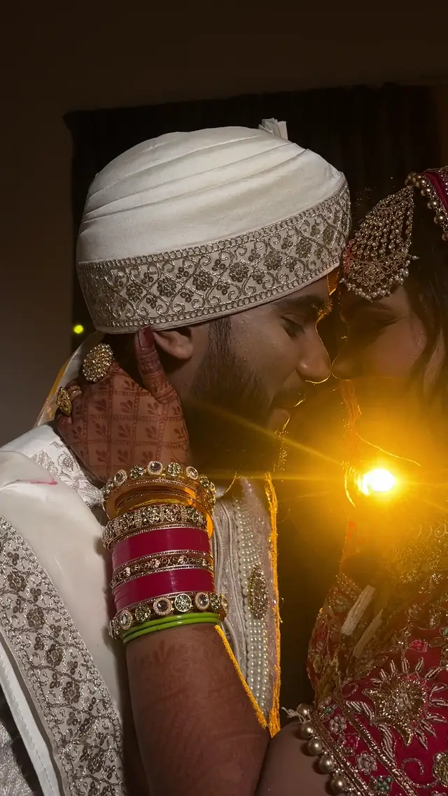 A groom in a cream turban leaning close to the bride, her bangled hand between them and a warm light flaring behind.