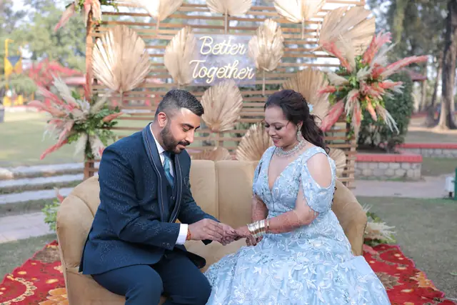 A groom in a navy suit putting a ring on the bride's finger, the two of them seated on a sofa on a lawn.
