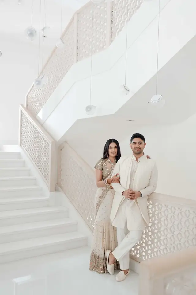A couple in ivory and gold standing against a white perforated staircase.