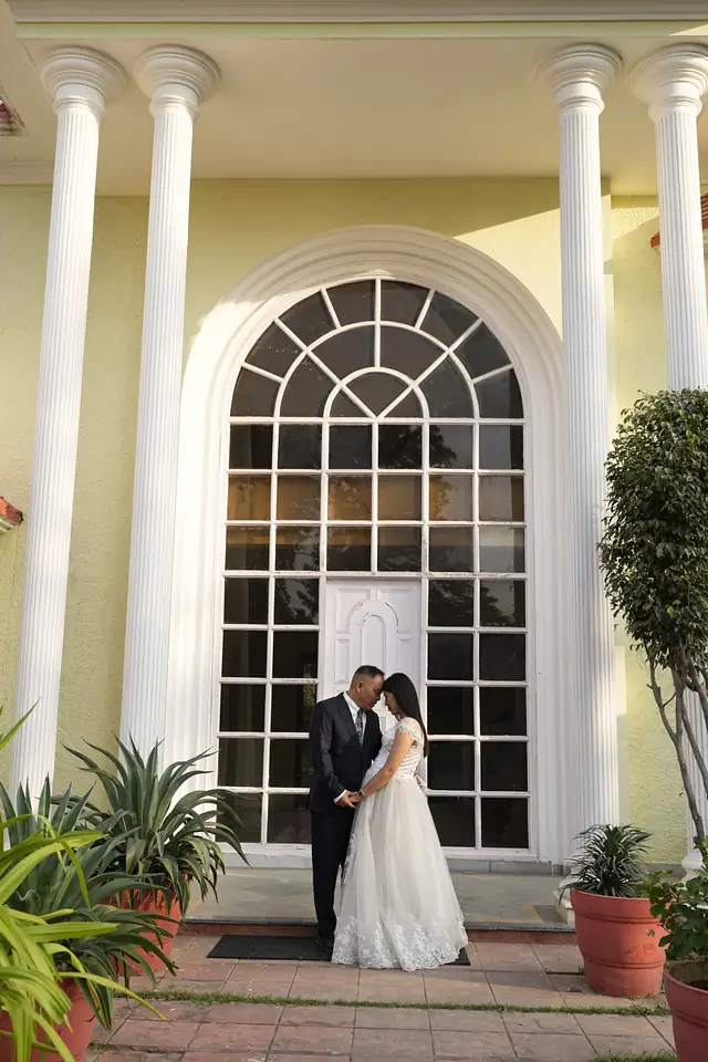 A couple in front of a tall arched window framed by white columns, potted plants on either side.